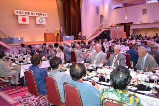 食事を前に高齢者たちが談笑しながらステージに注目しており、和やかな雰囲気の中で敬老会が進行している写真