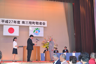 舞台の上でスーツ姿の男性が高齢の男性に向けて表彰を行っており、周囲には関係者や司会者が立って見守っている写真