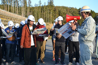 メガホンを使って説明している作業員の話を、見学者たちが資料を見ながら熱心に聞いている写真