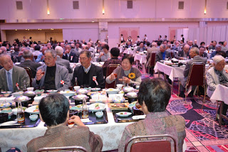 宴席で着物姿やスーツ姿の高齢者たちが料理を楽しみながら語らい、給仕係が奥で待機している広い会場の写真