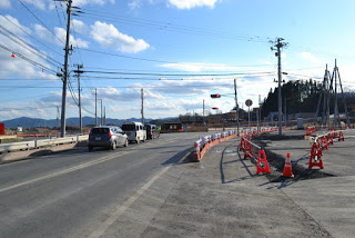 空は晴れて雲が少し浮かび、背景には建物や木々が広がり、道路沿いには電柱と電線が見え、左側には舗装された道路を走る2台の車があり、交通誘導のための三角コーンやバリケードが設置された道路工事現場の写真