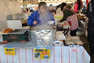 「焼ほや300円」「漁師はっと300円」「海鮮ごはん300円」などの値札が掲げられているイベントの販売ブースで、青いジャケットを着た女性スタッフがアルミホイルで覆われた大鍋から料理をよそっている様子の写真