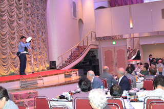 ステージ上で警察官のような衣装の出演者がパフォーマンスを披露し、観客の高齢者たちが注目している写真