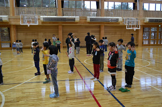 壁に向かって立っている子供たちと、ステージに向かって立っている子供たちが2チームに分かれて列を作って並び、発声練習を行っている写真