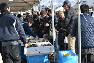 テントの下で多くの人々が列を作り、箱に入った商品を見たり購入したりしている、寒そうな屋外のにぎやかな福興市の様子の写真