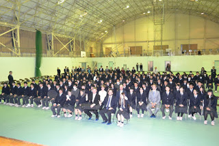 床には緑色のマットが敷かれ、上から照明が空間を照らされ、天井が高く金属の骨組みが見える広い体育館で、制服を着た大勢の生徒たちの間の椅子に演奏者がそれぞれ座っている記念写真