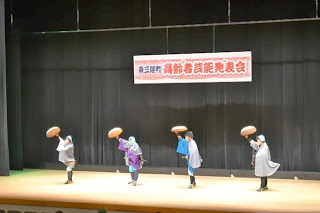 舞台の上で4人の出演者が和装で編み笠を頭の上に掲げて踊っており、背景には「南三陸町 高齢者芸能発表会」と書かれた横断幕がかかっている発表会の写真