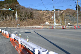 空は晴れていて昼間であることがうかがえ、背景には丘や木々、電柱と電線が見え、交通信号機とバリケードが設置された広くて空いている交差点の様子の写真