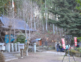 背の高い木々に囲まれた静かな屋外に、石灯籠と階段が神社へと続き、傾斜のある屋根を持つ伝統的な神社の前に人々が集まり、赤い旗が二本立てられている神社の写真