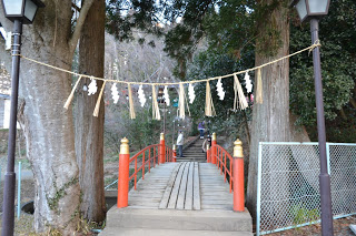 奥には神社へと続く石段が見える両側に大きな木が立ち、両側の街灯に白い紙垂がついた伝統的な注連縄が掛けられ、赤い欄干と金色の装飾が施された小さな木橋の入り口の写真