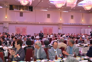 宴会場でテーブルを囲んだ高齢者たちが舞台に向かって拍手をしており、全体が明るい雰囲気で賑わっている写真