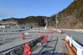 空は青く晴れていて、背景には丘の上の木々といくつかの建物が見え、白いガードレールに沿ってオレンジのバリケードで囲まれた道路があり、赤い交通バリケードと三角コーンが設置された工事現場の様子の写真