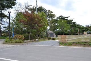曇り空の下、舗装された道路の奥に砂利道が続き、左側には街灯と赤い葉をつけた木々や大きな岩の記念碑があり、右側には白いフェンスが見える、自然豊かな上の山公園の写真