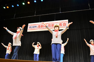 舞台上に揃いのセーラー帽と赤いスカーフを身につけた女性たちが笑顔で手を広げて踊っている様子の写真