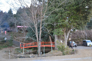 右側に数台の車が停まっている木々に囲まれた自然の中で、小さな朱色の橋が小川に架かっている境内の写真
