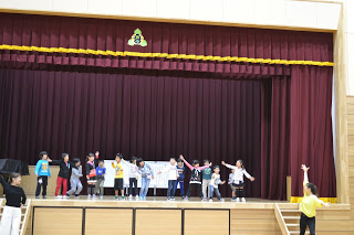 ステージ上で子どもたちが列になり、左右に腕を広げて演技をしている最中で、ステージの前に立った指導者が右手を高く挙げて一緒に踊っている様子の写真