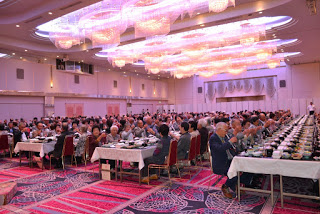 豪華なシャンデリアが輝く大きな会場で、多くの高齢者たちがテーブルに座りながら拍手をしている写真