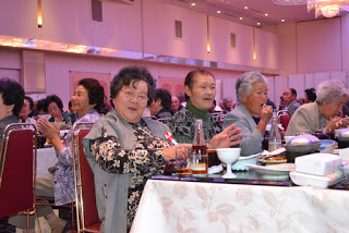 高齢の女性たちがテーブルに並べられた料理を前に楽しそうに会話しながら拍手している、笑顔あふれる様子の写真