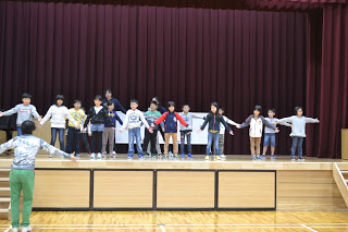 体育館のステージ下で指導している男性を見ながら、ステージ上で、子供たちが腕を広げて立ち同じような動きをしている練習の様子の写真