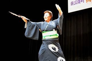 黒い着物に緑の帯を締めた女性が舞台上で扇子を片手に高く掲げ、もう一方の手を広げて華やかに踊っている写真