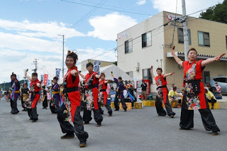 赤と黒の衣装を着た女性たちが腕を高く上げ、揃った振り付けで元気よく踊っている写真