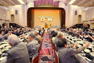 敬老会の開会式で舞台に立つ来賓に向かって、長テーブルに座る大勢の高齢者たちが一斉に杯を掲げて乾杯している場面の写真