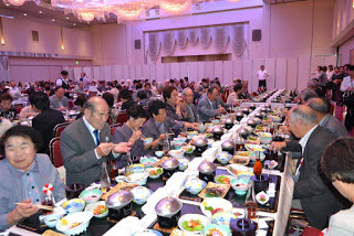 宴会場の中央に並んだ長いテーブルで、高齢者たちがにぎやかに料理を楽しんでいる写真