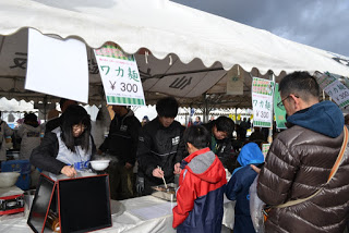 テントの上に「ワカ麺 300円」などのポップが掲げられた屋外のイベントや市場で、子どもや大人が並んでいる屋台の様子の写真