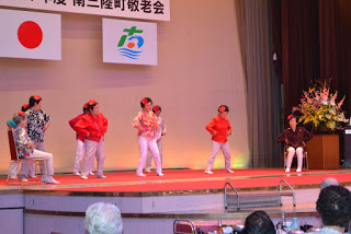 カラフルな衣装を着て頭には赤い花飾りをつけた人たちが腰に手を当てステージ上で元気よくダンスを披露している写真