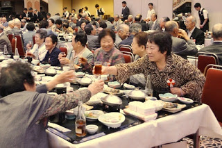 大広間の長テーブルで多くの高齢者が食事を楽しむ中、赤白のリボンをつけた女性が向かい側の参加者にグラスを差し出して乾杯しようとしているにぎやかな場面の写真