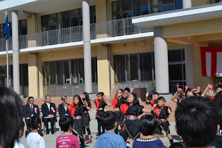 新しい校舎を背景に、赤と黒の衣装を着た児童たちが元気に踊っている様子を多くの来場者が見守っている運動場での発表の写真