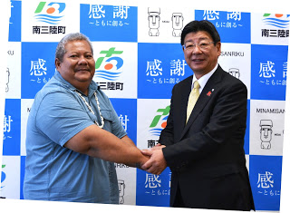 バックパネルの前で、スーツ姿の佐藤町長と水色のシャツを着たアダチ知事が握手をしている様子が写っている写真