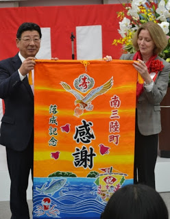 オレンジと黄色のグラデーション背景に、鷲や2つの島が浮かぶ海、魚やタコ、「祝」と書かれた宝船がデザインされ、「南三陸町 落成記念 感謝」と書かれた華やかな大漁旗を佐藤町長と左胸に赤い胸章リボンをつけた女性が持っている記念式典の写真