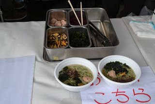 テーブルの上の使い捨て容器に、麺・シイタケ・ワカメなどが入った「ワカ麺」が2つ並び、その奥にはキノコや野菜、肉団子などの具材が仕切られた金属トレーと箸が置かれている写真