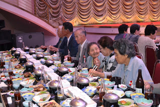 舞台前の長いテーブルにずらりと料理が並び、数人の高齢者が笑顔で食事を楽しんでいる写真