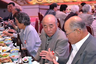 テーブルを囲んだ高齢の男性たちが食事をしながら会話をしており、親しげに話し込んでいる様子の写真