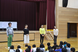 体育館の舞台の前で、4人の劇団四季の方々が正面を向いて立っており、前に座っている子どもたちへ話をしている様子の写真