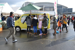 背景にはいくつものテントや屋台が並ぶ屋外の市場で、傍に傘が設置された黄色いフードトラックの周りに子どもや大人が並んでいる様子の写真