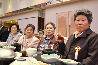 胸に赤白のリボンをつけた女性たち4人がカメラに向かって微笑みながら食事を楽しんでいる様子の写真