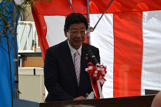 赤白の幕の前でスーツ姿の男性が胸にリボンを付けて演台に立ち、マイクでスピーチをしている写真