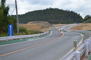 中央に白い区切り線が引かれたカーブした片側一車線の道路が、奥の森林地帯へと続いており、両側にはガードレールと工事資材が並び、左側には「仙台方面」と書かれた青い標識が立っていて、数台の車が走っている工事中の道路の写真