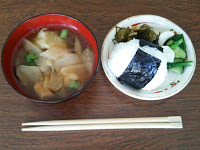 木の机の上に割り箸が置かれ、具だくさんの味噌汁、漬物が添えられたおにぎりが入ったお皿が並べられている写真