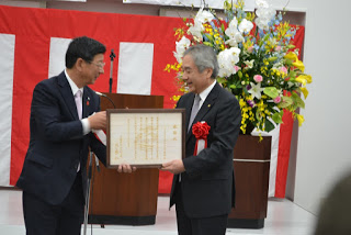 紅白幕が飾られ、大きな花の飾りが置かれている前で、佐藤町長が左胸に赤い胸章リボンとつけた男性に額縁に入った感謝状を渡している表彰式の写真