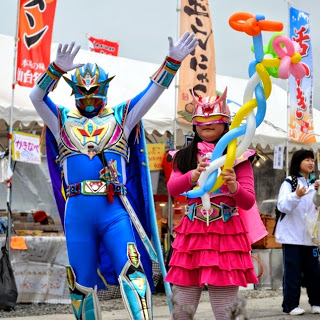 ヒーローのコスチュームを着た大人の男性と、ピンク色の戦士風衣装を着た女性が風船アートを持って祭りの屋台前でポーズをとっている写真