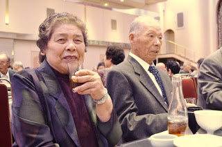 女性がグラスを手にして笑顔で乾杯のポーズをとり、隣の男性とともに食事の席で和やかに過ごしている様子の写真