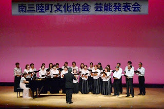 舞台上で中央に指揮者が立ち、左側の女性のピアノ演奏に合わせて、白と黒の衣装を着た大人たちが合唱を行っている様子の写真