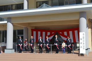 赤白の幕の前で、スーツ姿の大人と一人の子どもが並び、赤いリボン付きのはさみでテープカットをしている校舎前の式典の写真