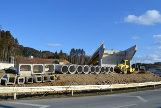 空は青く晴れて雲が少し浮かび、周囲には木々や丘が広がり、傾斜のある大きな建設中の橋があり、黄色の建設車両が見え、地面に複数のコンクリート製の管や構造物が並べられた工事現場の様子の写真