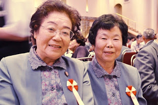 おそろいの服に赤白のリボンをつけた二人の女性が仲良く寄り添って笑顔を見せている様子の写真