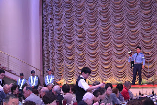 舞台の上に制服姿の警察官が立っており、テーブルでは高齢者たちが食事を続けており、ステージ左には警備員らしき人々も立っている写真